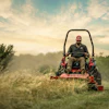 Red zero-turn lawn mower cutting grass at sunset with misty landscape background