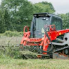 Red skid steer clearing vegetation in grassy field with trees in background
