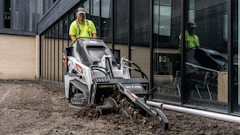 Worker operating Bobcat compact track loader for site preparation near modern building entrance