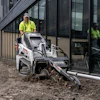 Worker operating Bobcat compact track loader for site preparation near modern building entrance