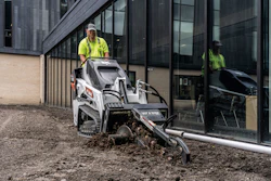 Worker operating Bobcat compact track loader for site preparation near modern building entrance