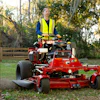 Worker operating red Ferris zero-turn mower in safety vest on grassy property with trees