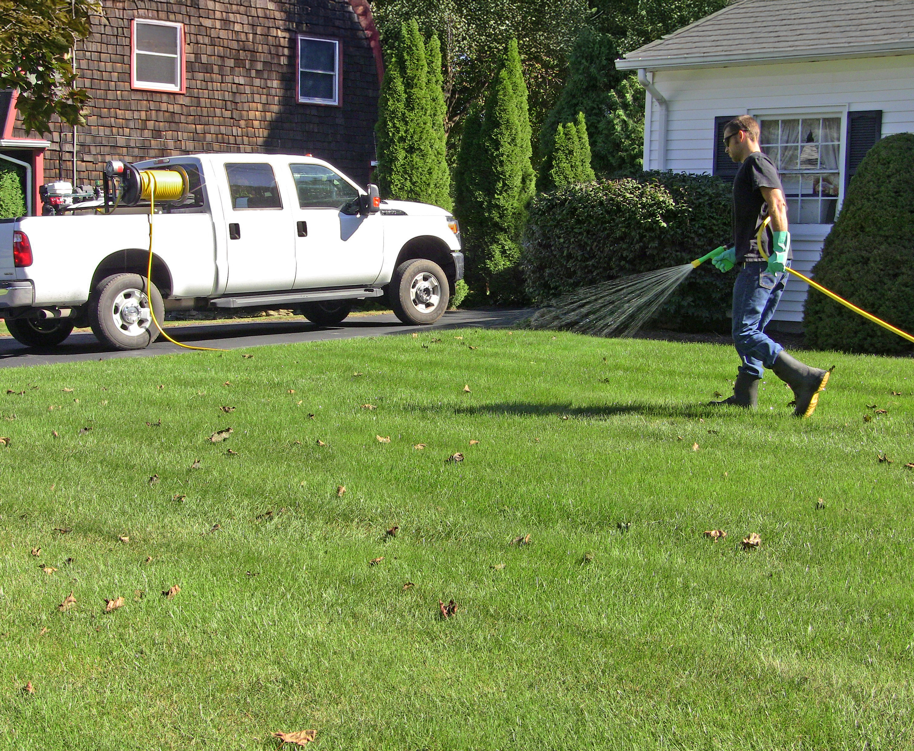 A lawncare technician sprays pesticides on a residential lawn using a hose reel sprayer.