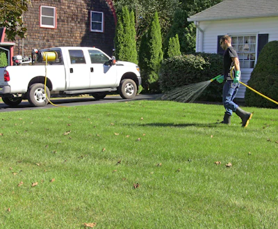 A lawncare technician sprays pesticides on a residential lawn using a hose reel sprayer.