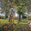 Person wearing protective mask and gloves raking leaves in a park with tall trees and green grass