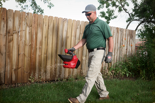 Man in green shirt carrying red yard waste container while walking on grass near wooden fence
