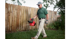 Man in green shirt carrying red yard waste container while walking on grass near wooden fence