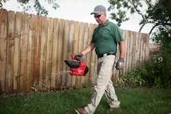 Man in green shirt carrying red yard waste container while walking on grass near wooden fence