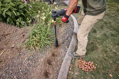 Person using a red and black string trimmer along a garden pathway with plants and grass visible