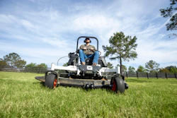 Operator on zero-turn radius mower cutting grass in open field with trees and fencing