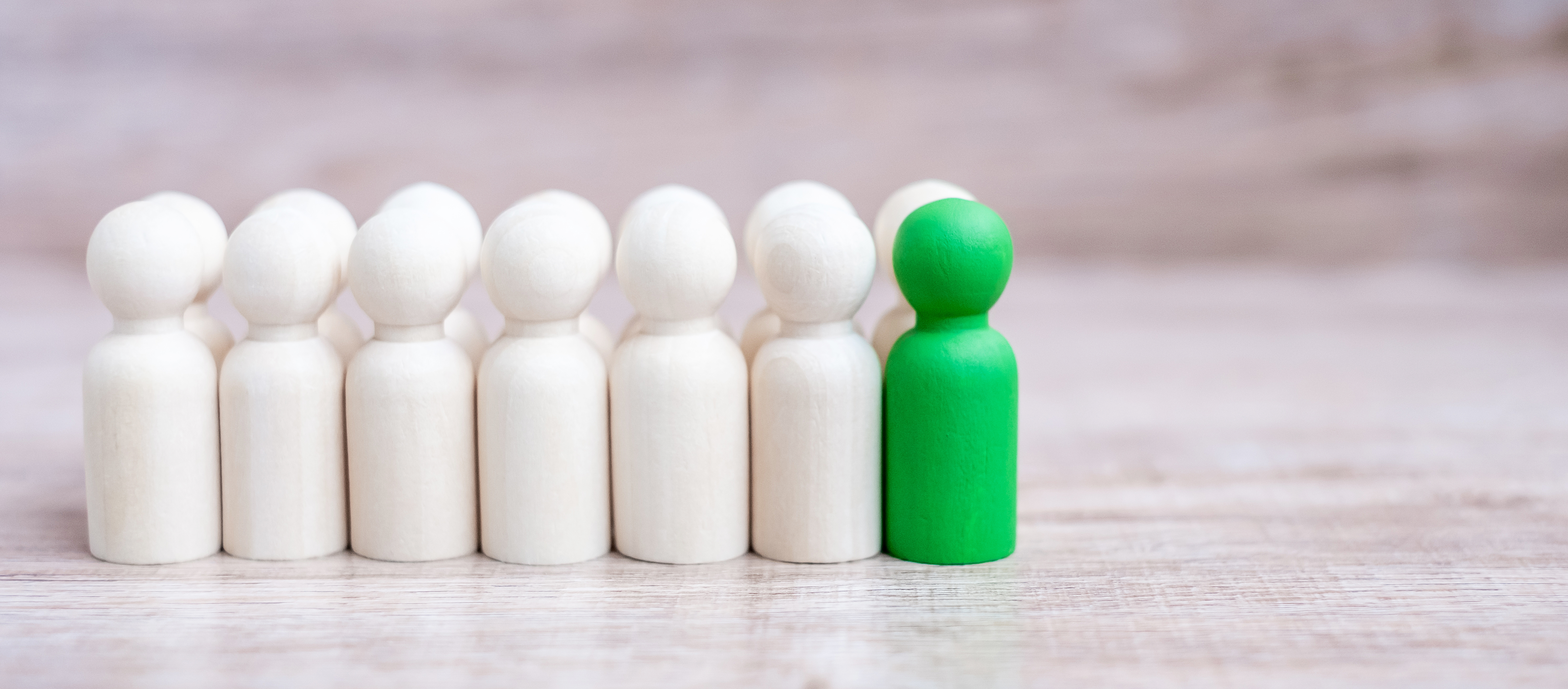 Green peg doll standing out among six white peg dolls on wooden surface