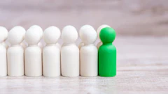 Green peg doll standing out among six white peg dolls on wooden surface