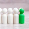 Green peg doll standing out among six white peg dolls on wooden surface