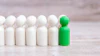 Green peg doll standing out among six white peg dolls on wooden surface