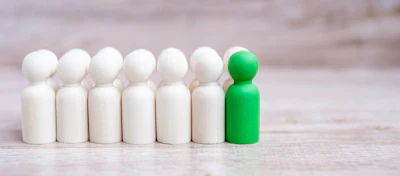 Green peg doll standing out among six white peg dolls on wooden surface