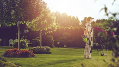 Person standing in sunlit garden with manicured lawn, flowering plants, and tall trees during golden hour
