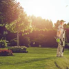 Person standing in sunlit garden with manicured lawn, flowering plants, and tall trees during golden hour