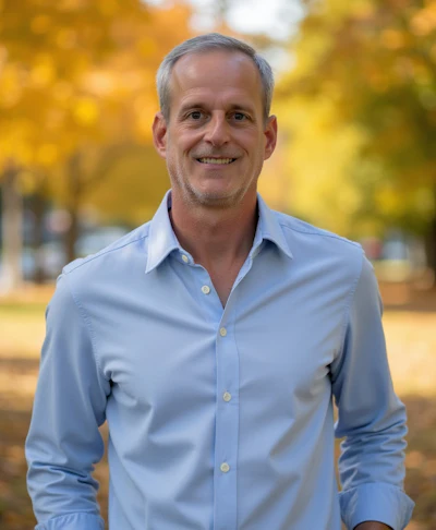 Professional headshot of a man in light blue button-up shirt smiling outdoors with autumn foliage background