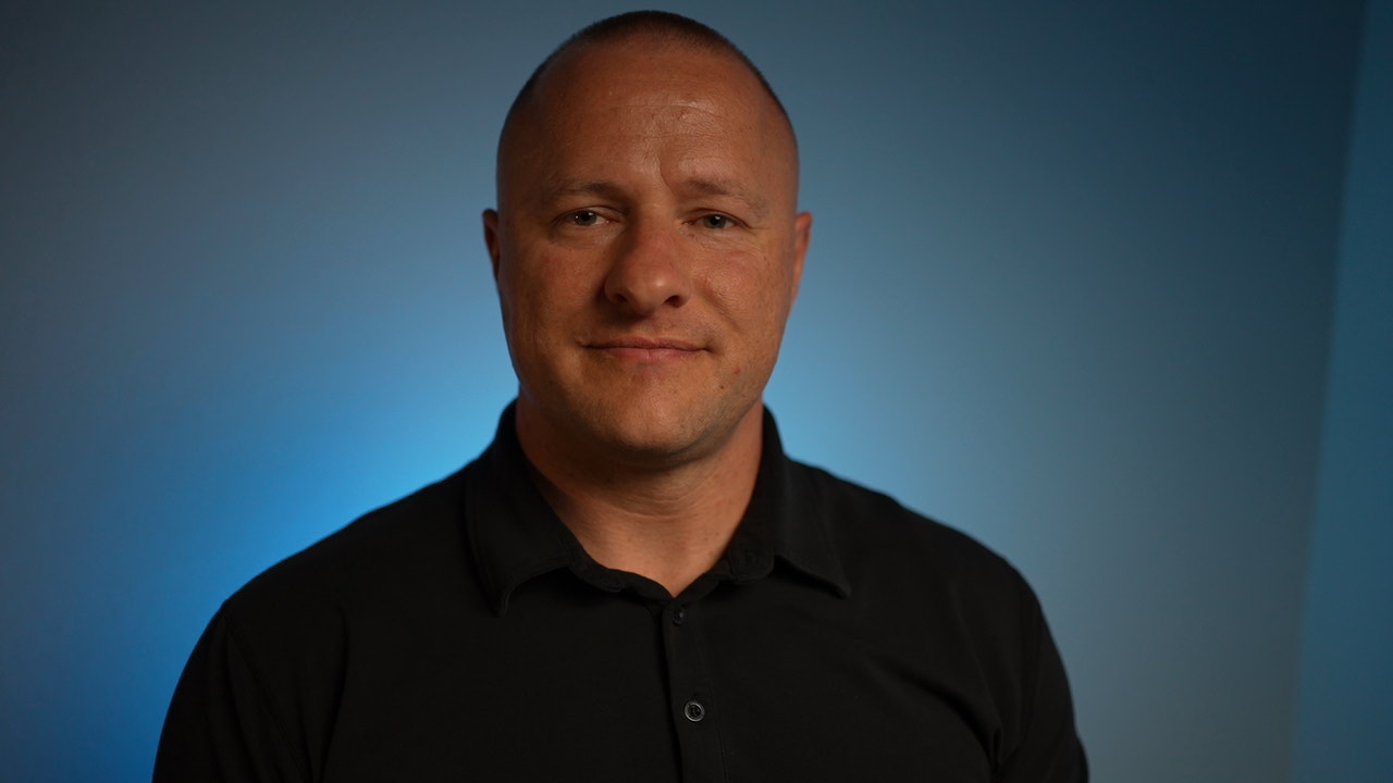 Professional headshot of a man wearing a black polo shirt against a blue gradient background