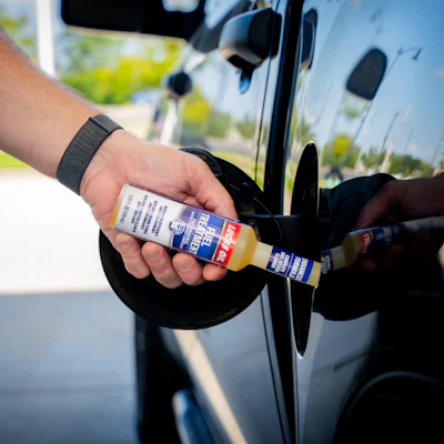 Person holding blue and white branded product bottle next to white vehicle door outdoors