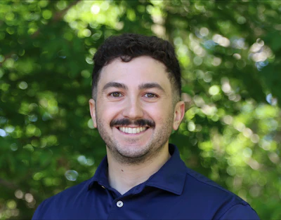 Smiling man with dark hair and mustache wearing navy polo shirt against green foliage background