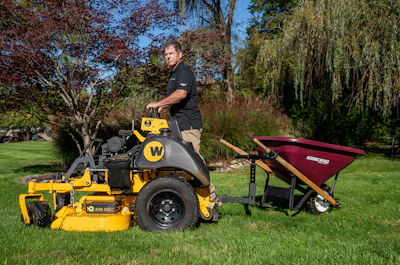 Man operating yellow zero-turn mower with attached maroon trailer in landscaped yard with trees