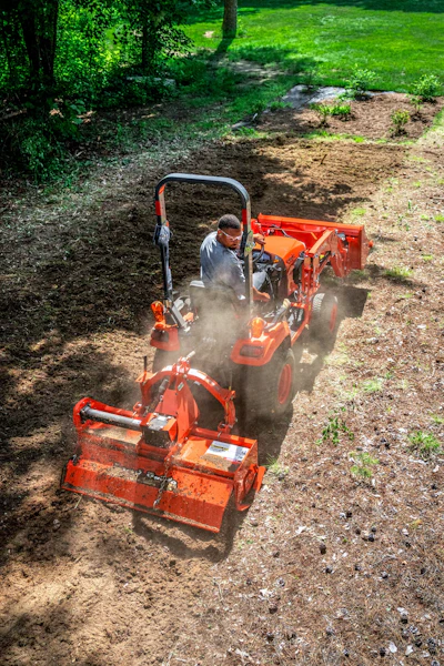 Operator using red tiller machine to cultivate garden soil with dust clouds rising, surrounded by green lawn and trees.