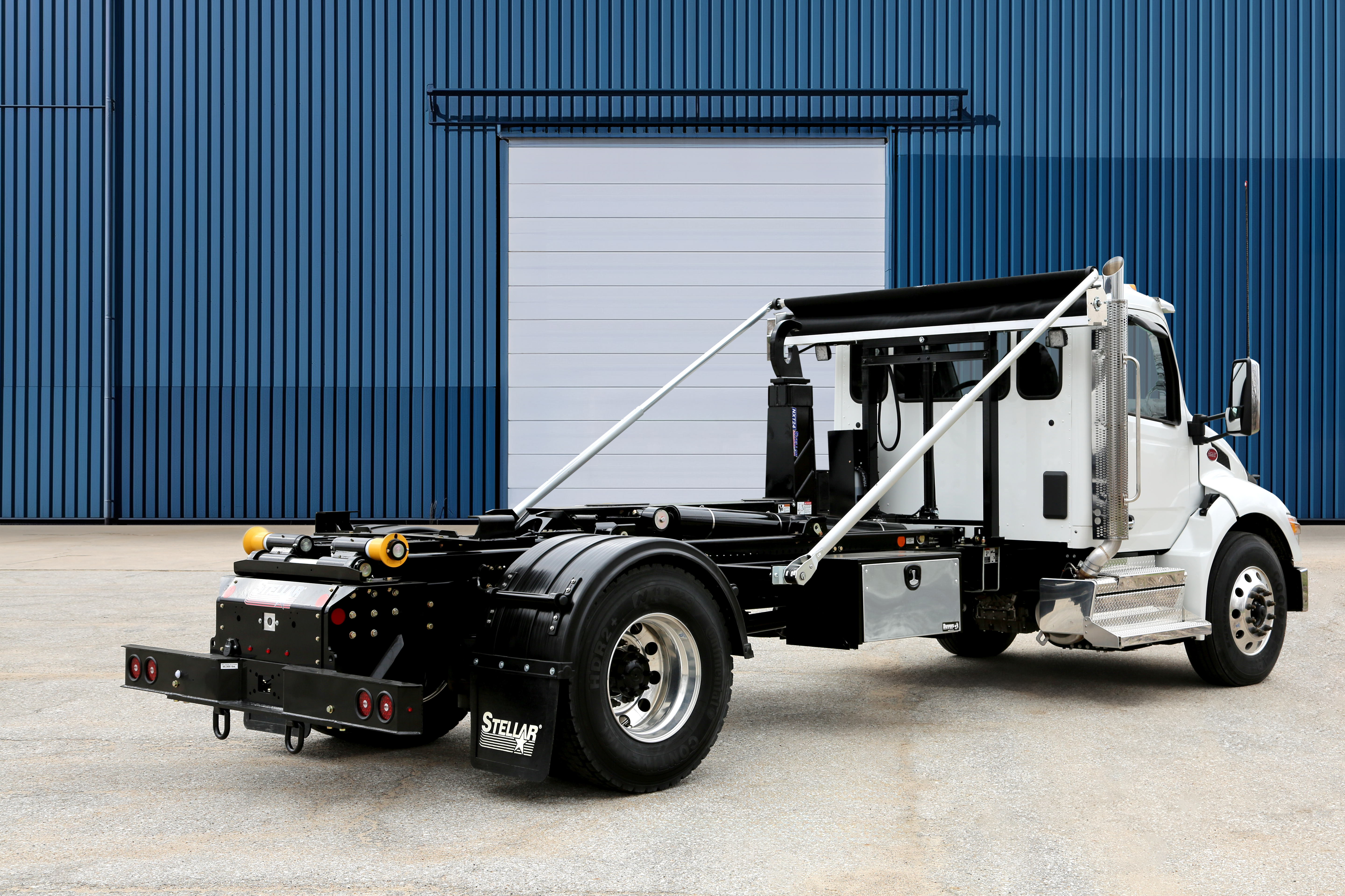 White and black commercial tow truck with hydraulic lift system parked in front of industrial blue corrugated metal building