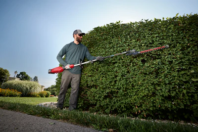 Man trimming tall green hedge with electric hedge trimmer in residential garden