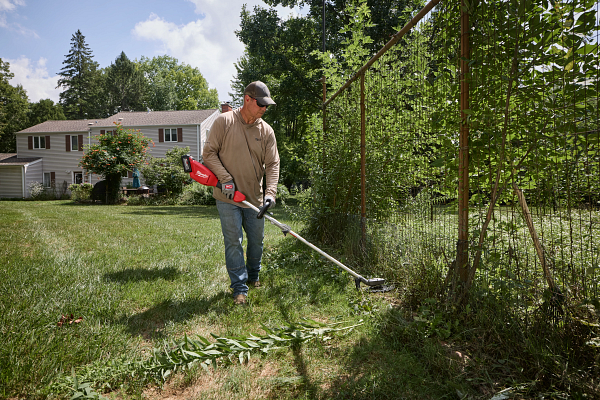Man using a red string trimmer to edge grass along a fence in a residential yard