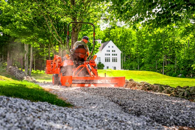 Orange wood chipper machine processing debris on gravel driveway with white house and forest background