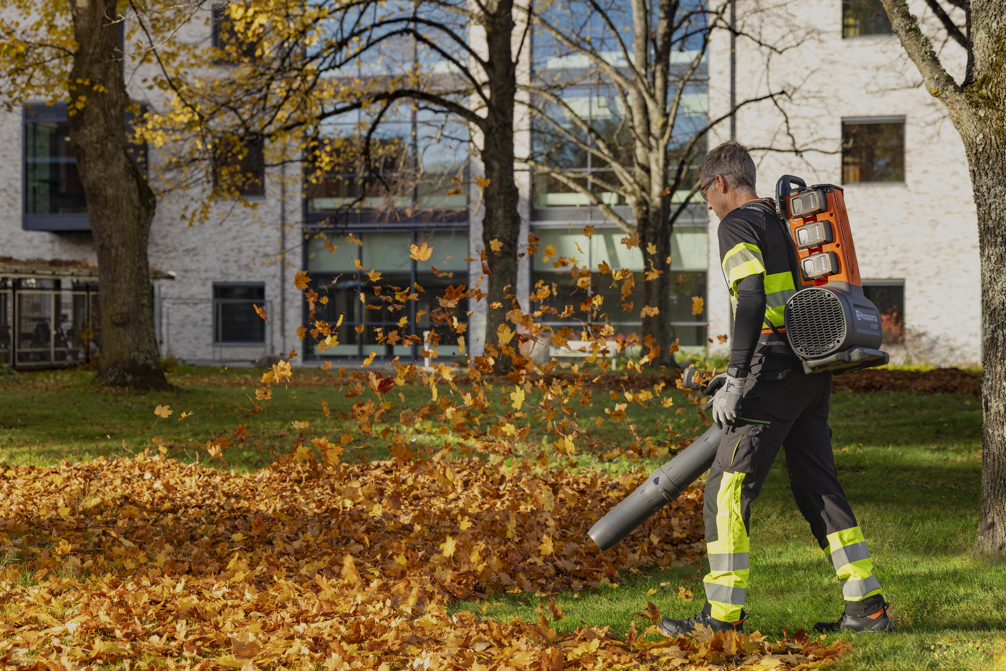Worker in safety gear using leaf blower to clear fallen autumn leaves from residential yard