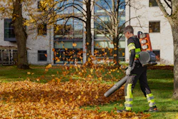 Worker in safety gear using leaf blower to clear fallen autumn leaves from residential yard