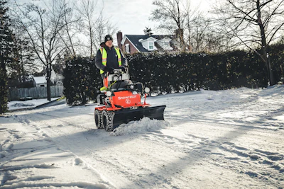Worker operating red snow plow equipment on residential street lined with snow and bare trees