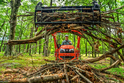 Operator on red Kubota tractor loaded with firewood in a dense forest surrounded by fallen logs