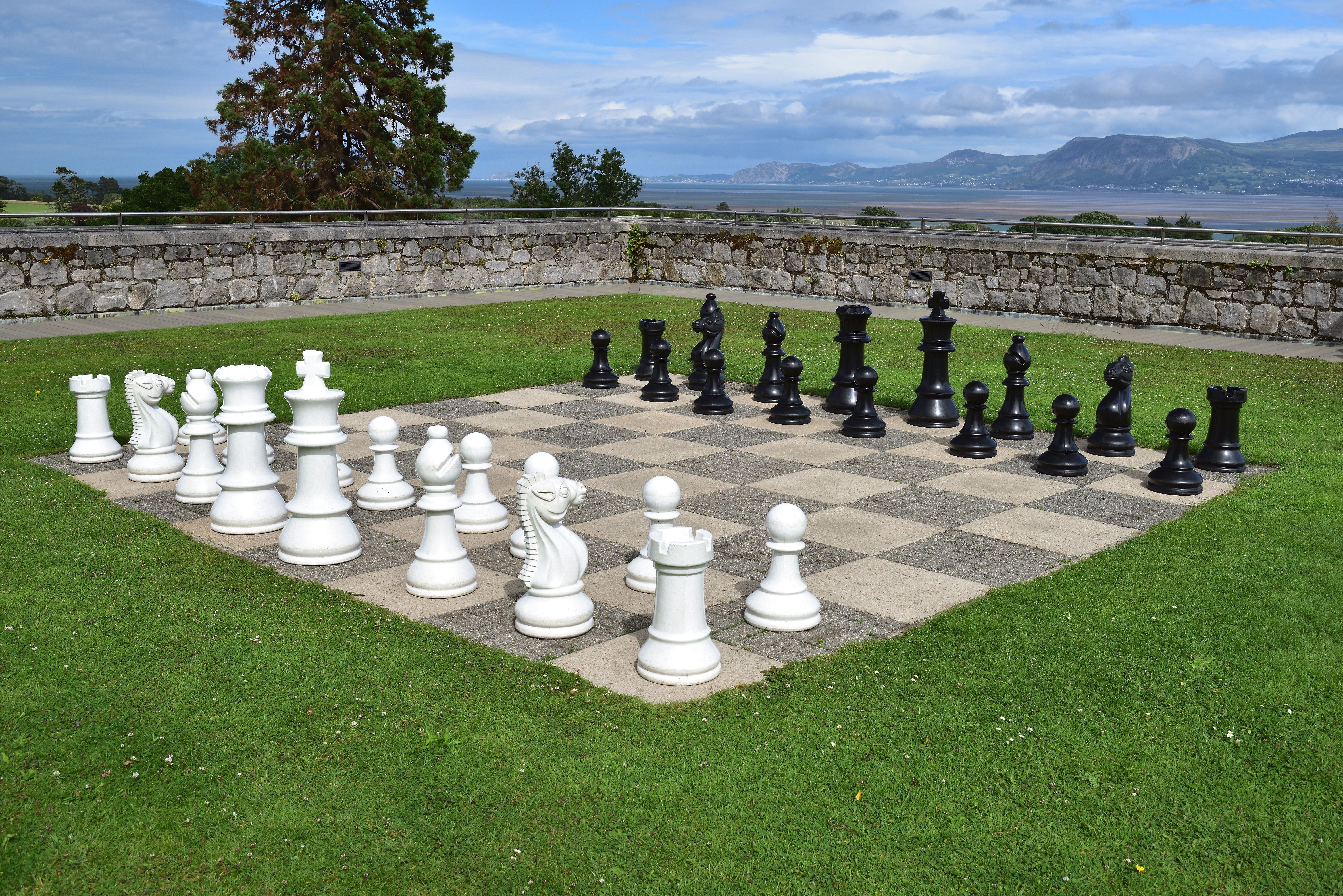 Outdoor giant chess game with white and black pieces on stone board in scenic lakeside garden setting