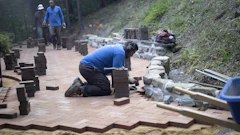 Workers installing brick pavers and stone materials on an outdoor patio pathway in a wooded area