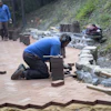 Workers installing brick pavers and stone materials on an outdoor patio pathway in a wooded area