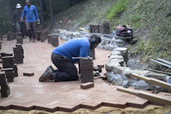 Workers installing brick pavers and stone materials on an outdoor patio pathway in a wooded area