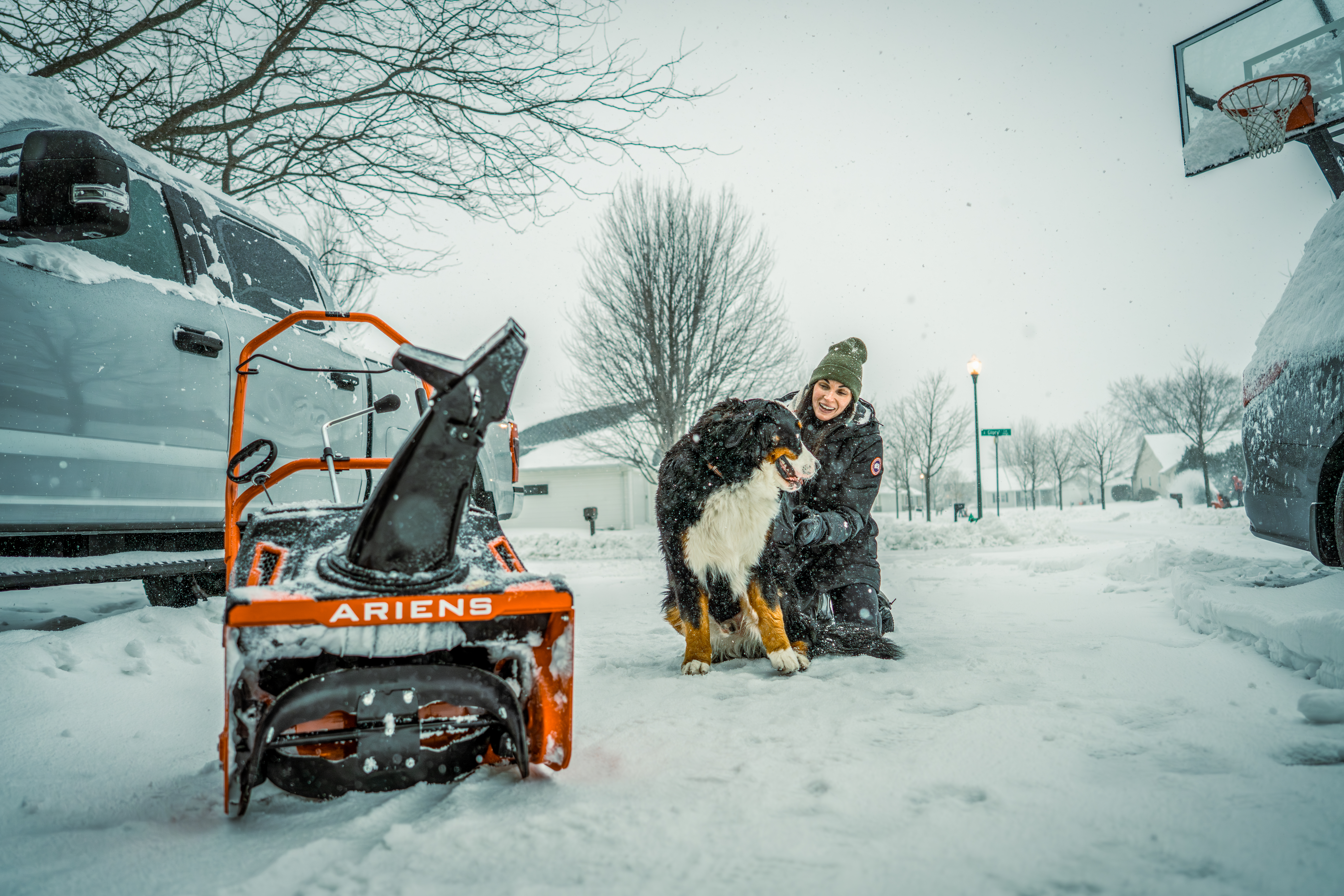 Person with dog beside orange Ariens snow blower during winter snowfall in residential area