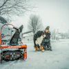 Person with dog beside orange Ariens snow blower during winter snowfall in residential area