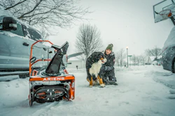 Person with dog beside orange Ariens snow blower during winter snowfall in residential area