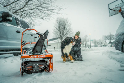 Person with dog beside orange Ariens snow blower during winter snowfall in residential area