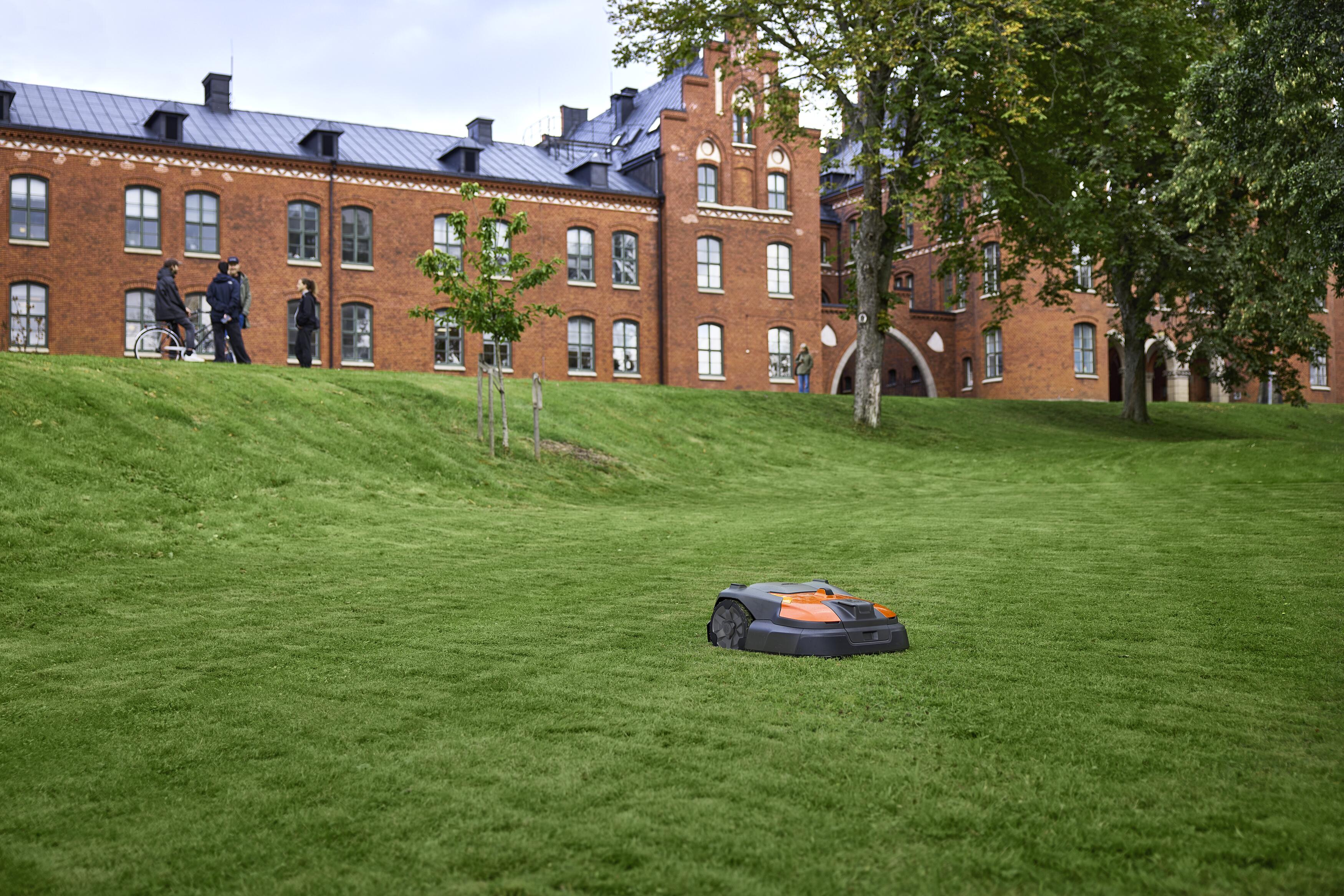 Autonomous robotic lawn mower operating on a sloped grass field in front of a brick university building with trees