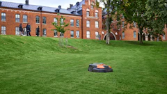 Autonomous robotic lawn mower operating on a sloped grass field in front of a brick university building with trees