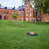 Autonomous robotic lawn mower operating on a sloped grass field in front of a brick university building with trees