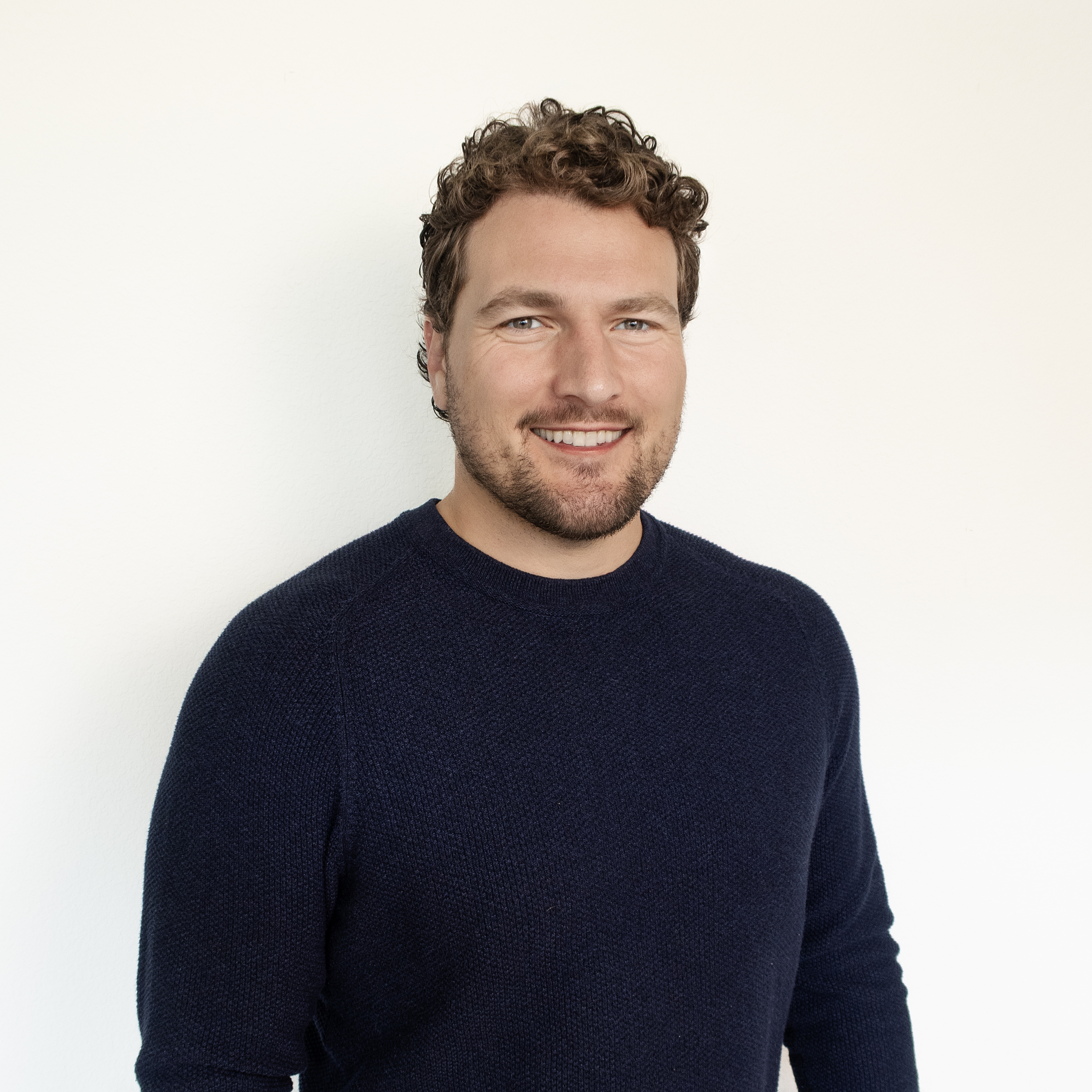 Professional headshot of a smiling man with curly brown hair wearing a dark navy sweater against a white background