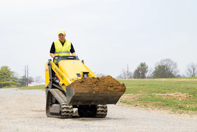 Construction worker operating yellow Bobcat skid steer loader with dirt bucket on rural road