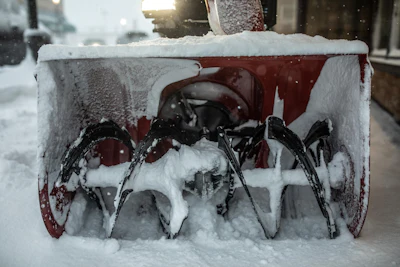 Snow-covered red and black snow blower parked on snowy street during winter storm