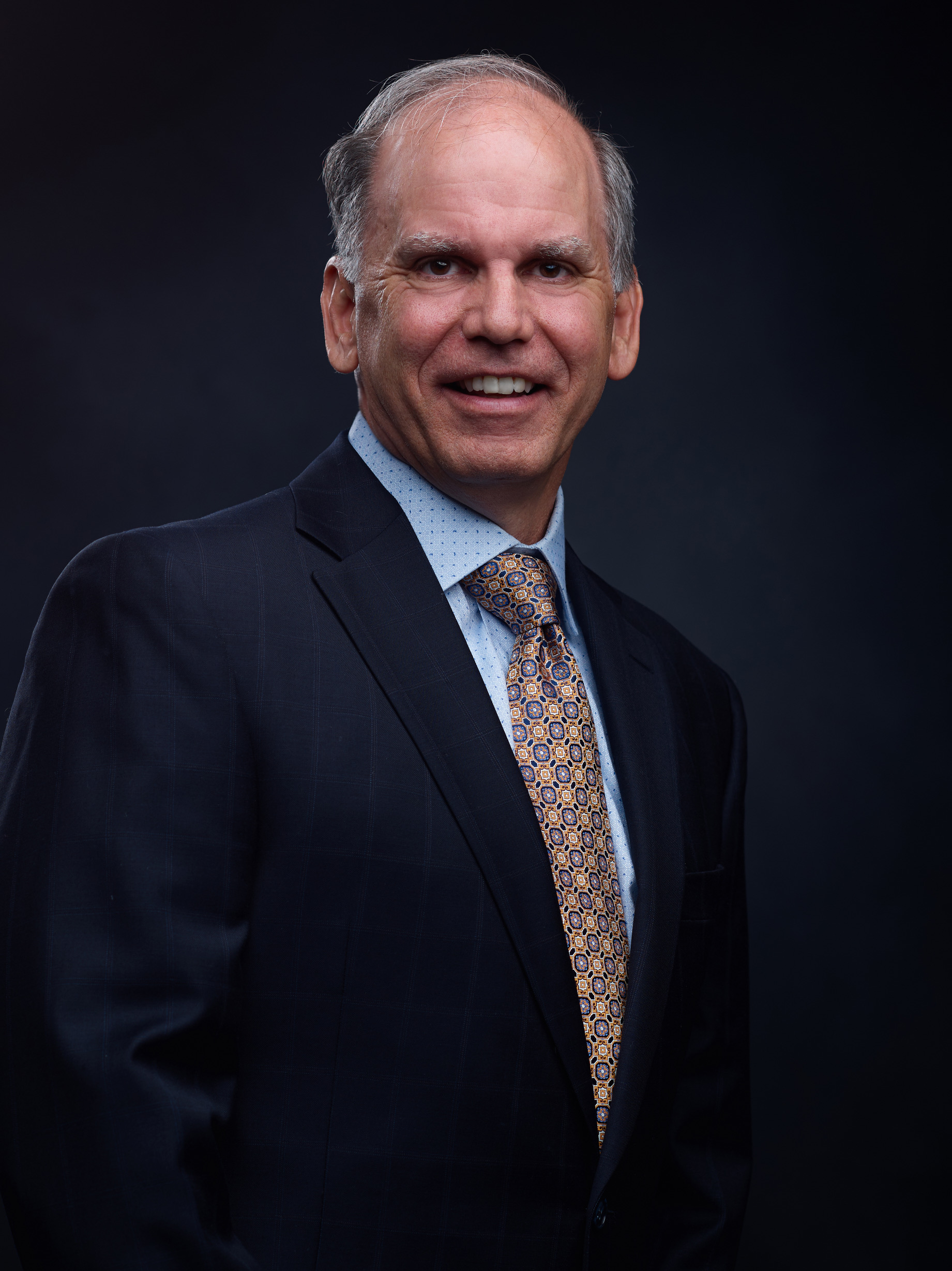 Professional headshot of a man in dark suit and patterned tie smiling against dark background Headshot